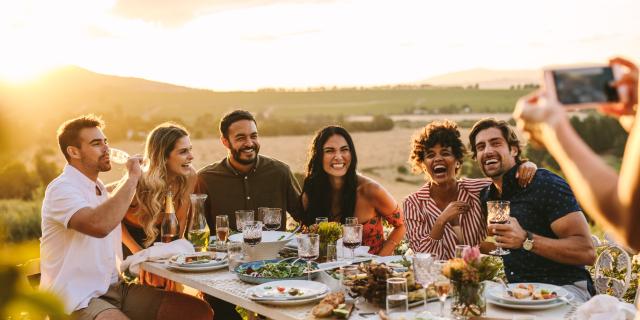 Group of men and women posing for a photograph during an outdoor dinner party. Woman hands taking picture of her friends at dinner party.